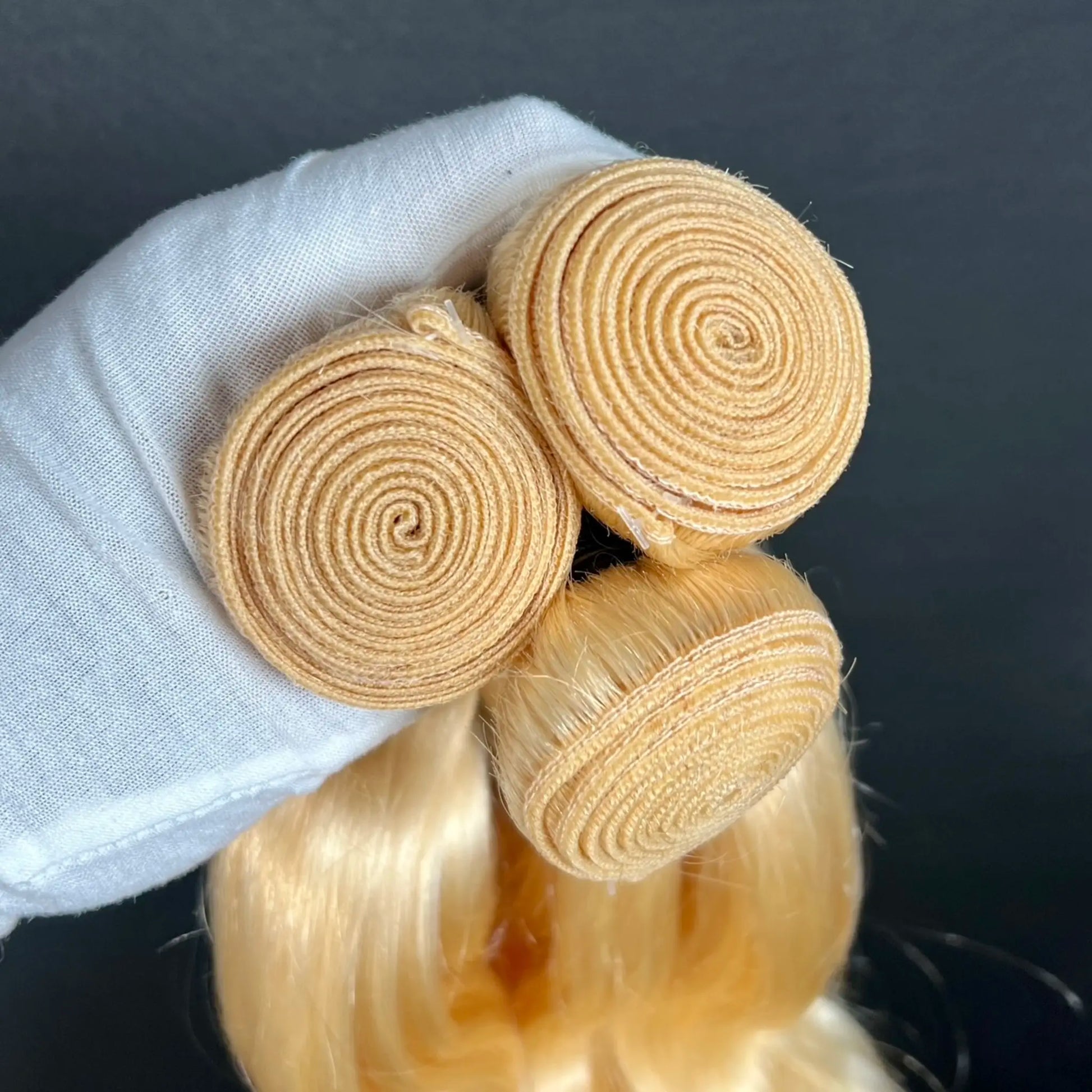 Three blonde hair extensions held by a gloved hand against a dark background