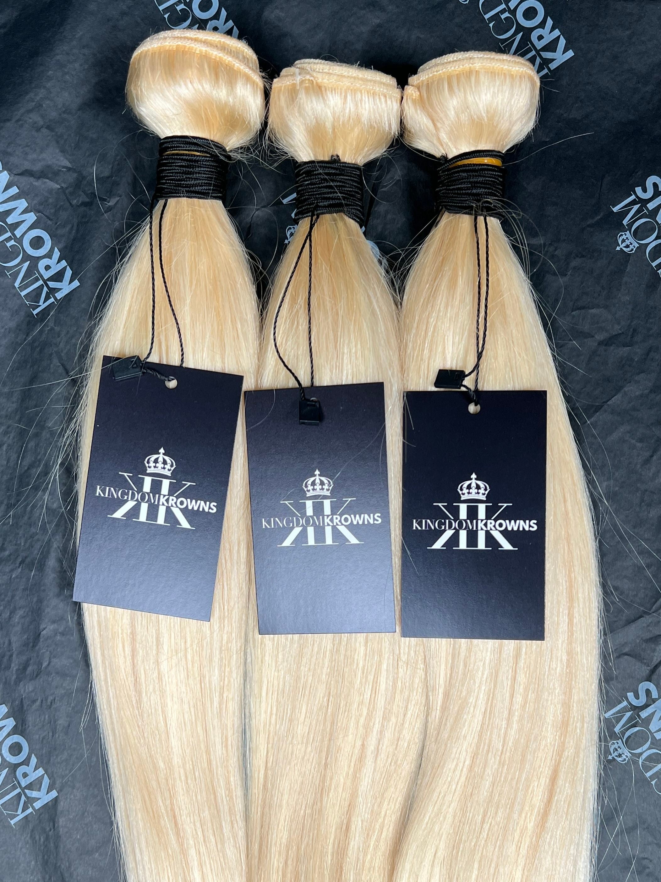 Three bundles of blonde hair extensions with branded tags on a dark background.